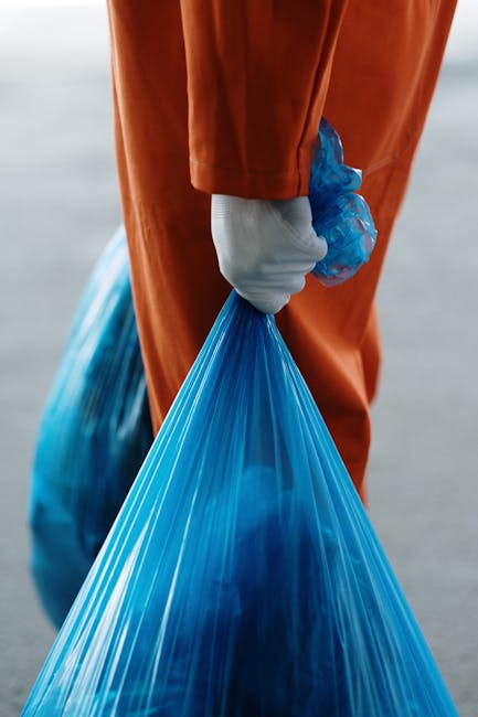 A person wearing an orange protective suit and white gloves is holding a large, partially filled blue plastic rubbish bag by its gathered top. The bag appears to contain waste material, with its trans